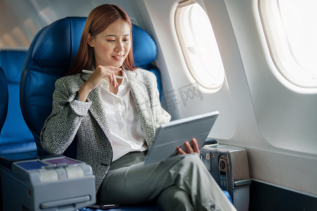 一位成功的亚洲女商人或女性企业家在飞机上穿着正式西装的肖像坐在商务舱的座位上，并在飞行过程中使用平板电脑