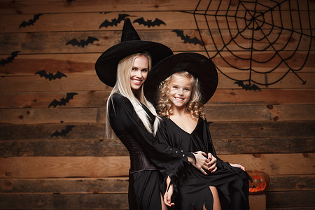 万圣节概念 — 快乐的母亲和她的女儿穿着女巫服装庆祝万圣节，在木制工作室的背景下，在蝙蝠和蜘蛛网上摆着弯曲的南瓜。