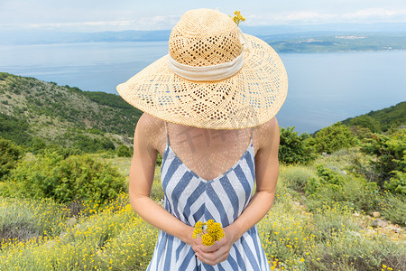 穿着条纹夏装、头戴草帽的年轻女子站在野花盛开的地方，在克罗地亚亚得里亚海沿岸美丽的大自然中捧着一束黄色花