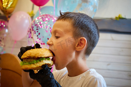 吃汉堡的男孩摄影照片_英俊的小男孩戴着黑色橡胶手套吃汉堡。