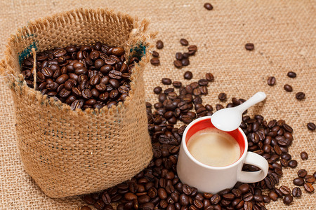 咖啡豆跌落摄影照片_帆布袋中的烘焙咖啡豆和一杯咖啡。