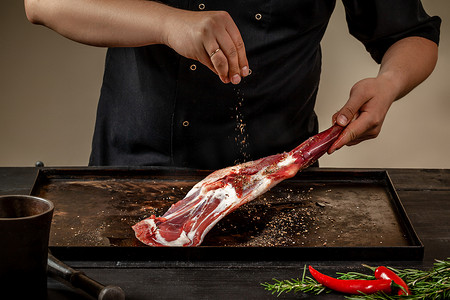 蒸羊腿摄影照片_男厨师在木桌上的石盘上用盐和胡椒摩擦生羊腿。