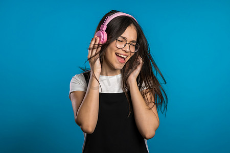 粉红kt板摄影照片_年轻漂亮的长发女孩在蓝色背景的工作室里玩得很开心，微笑着，戴着耳机跳舞。