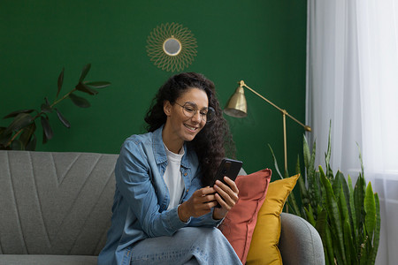 绿色的墙壁摄影照片_年轻美丽的西班牙裔女性在家坐在客厅的沙发上，客厅里有绿色的墙壁，