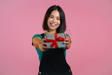 兴奋的女人拿着礼品盒，用手把它交给粉色墙壁背景上的相机。