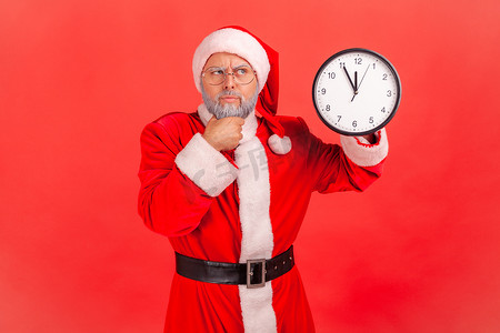 沉思的老人，留着灰胡子，穿着圣诞老人服装，站着，拿着挂钟，托着下巴，等待圣诞晚会，目光移开。