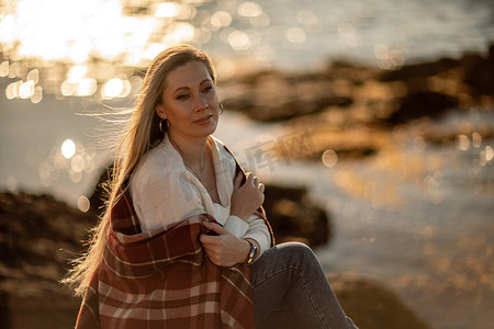 白毯子摄影照片_迷人的金发白种女人在日落时在海滩上享受时光，坐在毯子里，看着旁边，背景是日落的天空和大海。