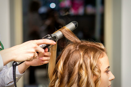 美容机摄影照片_理发师用卷发器为美容院里长着棕色长发的年轻女子做卷发。