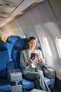一位成功的亚洲女商人或女性企业家在飞机上穿着正式西装的肖像坐在商务舱的座位上，并在飞行期间使用智能手机