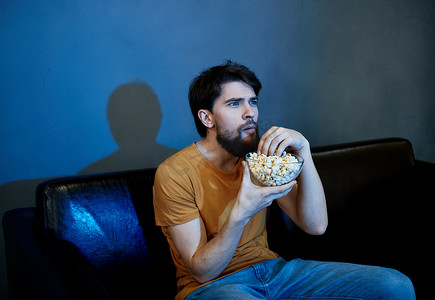 黑暗房间摄影照片_孤独的男人坐在沙发上，手里拿着爆米花盘灰墙
