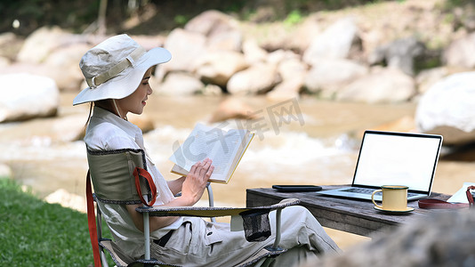 平静的女旅行者读书，享受在河岸附近独自露营。