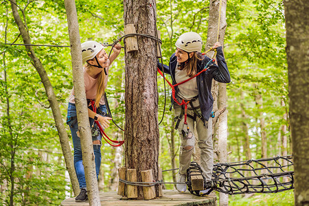 滑索摄影照片_两名女女孩在森林中的极端公路电车滑索中滑翔攀登，在树上的登山绳安全链接上到树顶绳索冒险公园。