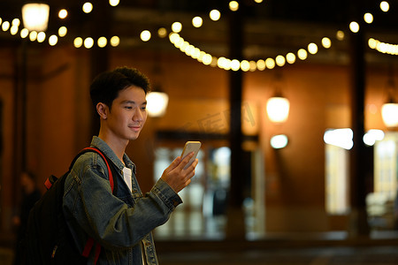 微笑的亚洲男子站在夜间路灯背景模糊的城市大道上检查手机