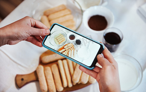 手使用智能手机相机，在白色桌布背景上拍摄提拉米苏甜点的配料照片。