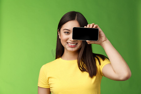 特写开朗乐观的亚洲女性推广智能手机应用程序，手持电话近眼注视相机，满意地微笑着介绍游戏，酷炫的新应用程序，站在绿色背景上