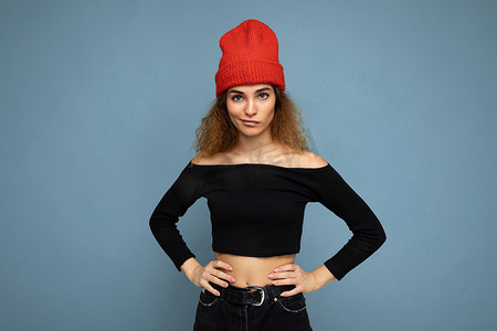 照片中，漂亮迷人、酷炫的年轻卷发深色金发女郎孤立地站在蓝色背景墙上，上面有文字复制空间，穿着日常时尚的黑色上衣和红色帽子