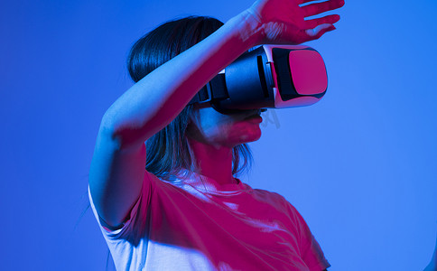 身穿白色 T 恤、兴奋的黑发女性使用虚拟现实 VR 耳机眼镜获得经验，在霓虹灯下双手打手势。