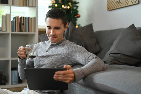 英俊的男人拿着咖啡杯，在家里用数字平板电脑上网。