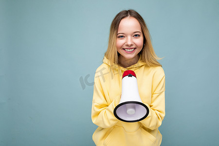 衫摄影照片_照片肖像美丽迷人的积极快乐的微笑金发年轻女子与真诚的情感穿着时尚的黄色连帽衫隔离在蓝色背景与复制空间和对扩音器说话。