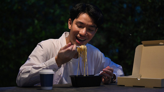 快乐的年轻亚洲男子在夜市的餐桌上吃外卖面条。