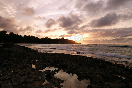 日落穿过云层，反射在海浪上，当它们冲破卡维拉湾潮汐池的岩石海岸时