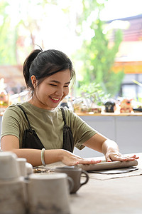 陶瓷工艺摄影照片_微笑的女人用擀面杖做粘土，在桌子上制作陶瓷，在车间制作手工陶瓷