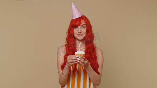 快乐的红发女孩庆祝生日周年纪念派对，在小蛋糕纸杯蛋糕上吹蜡烛