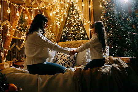 家庭环境摄影照片_一个小女孩和她的母亲在圣诞树旁边的沙发上享受舒适的家庭环境。