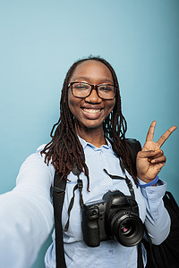 快乐微笑的摄影师与现代相机设备在自拍时给出和平标志。