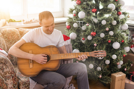 冬天的音乐背景摄影照片_年轻帅哥坐在沙发上，他正在弹吉他，音乐，圣诞节，放松概念