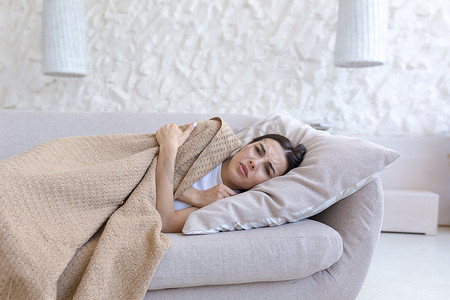 生病的年轻女子躺在家里沙发上的枕头上，身上盖着棕色毯子