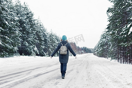 一个穿着灰色外套、戴着蓝色针织帽、背着灰色背包的女孩沿着冬季路边行走