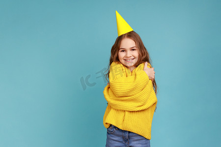 派对锥体中的小女孩拥抱自己，庆祝她的生日，看着镜头微笑。