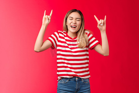 酷炫派对摄影照片_乐观狂野鲁莽的金发亚洲女孩玩得失去控制，展示摇滚标志享受很棒的派对，在酷炫的音乐会上大喊大叫，重金属标志闭上眼睛，站在红色背景