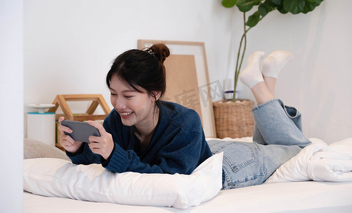 早上，在卧室的白色床垫上画出美丽的年轻亚洲女性的肖像。智能手机掌握在女性手中。