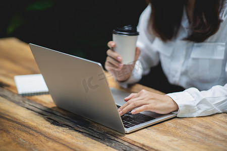 一杯热咖啡摄影照片_特写手的女商人在笔记本电脑上工作，并在木桌上拿着一杯热咖啡和一杯咖啡。