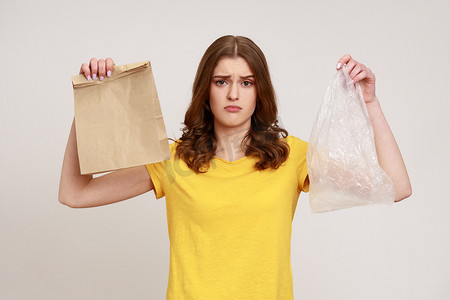 两分类摄影照片_穿着黄色休闲 T 恤的年轻心烦意乱的少女手里拿着两个袋子，纸袋和塑料袋，看着镜头悲伤，拒绝使用塑料。