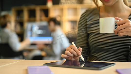 年轻创意女性拿着咖啡杯并使用数字平板电脑在工作场所的剪影