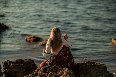 迷人的金发白种女人在日落时在海滩上享受时光，坐在毯子里，看着旁边，背景是日落的天空和大海。