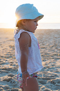 戴着巴拿马帽的金发小女孩站在海滩上，看着大海