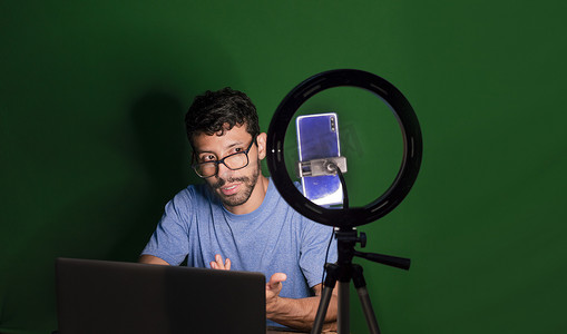 手持铃摄影照片_用笔记本电脑制作视频博客的年轻人，用手机制作内容的年轻人创作者，用笔记本电脑进行绿色背景视频通话的男人，