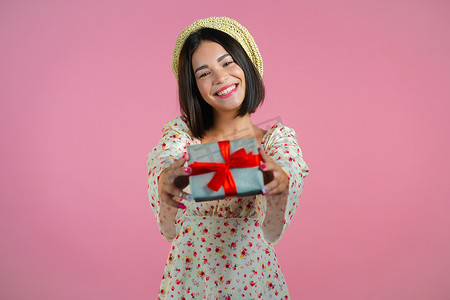 兴奋的女人拿着礼品盒，用手把它交给粉色墙壁背景上的相机。