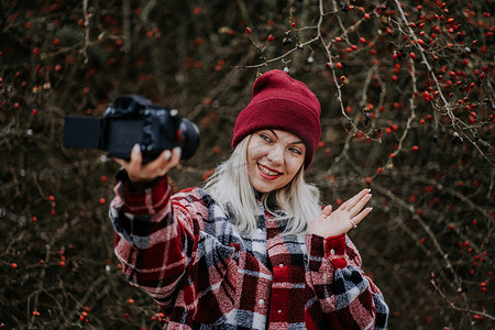 旅游博客摄影照片_微笑的旅游博主女士用她的相机在自然森林背景下制作新的视频博客、视频博客。