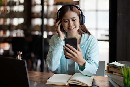 戴着无线耳机的快乐亚洲女学生使用手机上的学习应用程序、在智能手机上观看网络研讨会、听音频课程、在打开的书本上学习、在家抄书