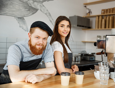 咖啡经营理念-积极的年轻留着胡子的男人和美丽迷人的女咖啡师夫妇穿着围裙，站在酒吧库特准备在现代咖啡店提供咖啡服务