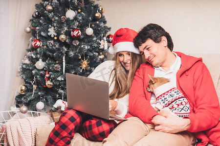 可爱的情侣夫妻的肖像，戴着红色圣诞帽的笔记本电脑，与穿着毛衣的小狗进行视频通话，在家享受圣诞节时光。