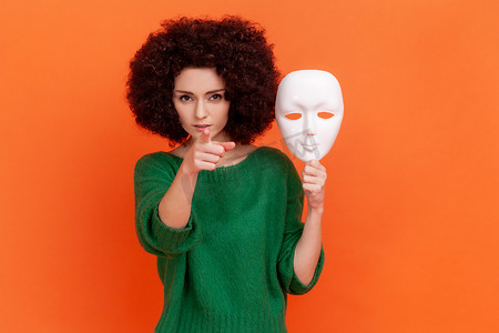 专横的摄影照片_严格专横的女人，留着非洲发型，穿着绿色休闲风格的毛衣，手里拿着白色面具，用手指着镜头，表情严肃。
