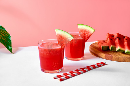 粉色夏天背景摄影照片_新鲜的西瓜汁或冰沙在玻璃杯中，粉红色背景上有西瓜片。