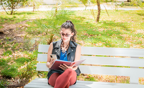 一个女孩坐着用笔记本和手机做笔记，女人坐着用手机做笔记，拉丁女孩在公园里用手机做笔记
