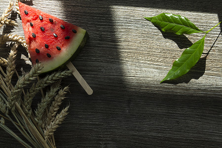 太阳表面摄影照片_冰淇淋架上放着一块三角形的甜西瓜，木质表面上放着一杯加冰的冷饮和成熟的麦穗。